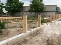 Solidny fundament pod ogrodzenie z drewnianymi słupkami, w tle budowa domu.