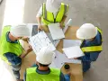 Zespół budowlańców w kamizelkach odblaskowych analizuje plany. Dyskusja o projekcie, prąd budowlany w tle.