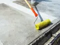 Wałek z żółtym włosiem rozprowadza środek do impregnacji betonu na szarej powierzchni.