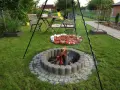 Ognisko w kamiennym kręgu, nad którym wisi okrągła taca z mięsem i warzywami na szaszłykach, gotowe do grillowania w ogrodzie.
