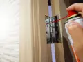 Dłoń z puszką sprayu wykonuje regulację zawiasów drzwi, aplikując środek smarny na metalowy element.