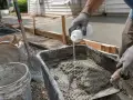 Rękawiczki i łopatka mieszają szybki beton w wózku. Obok leży worek z cementem i wiadro.