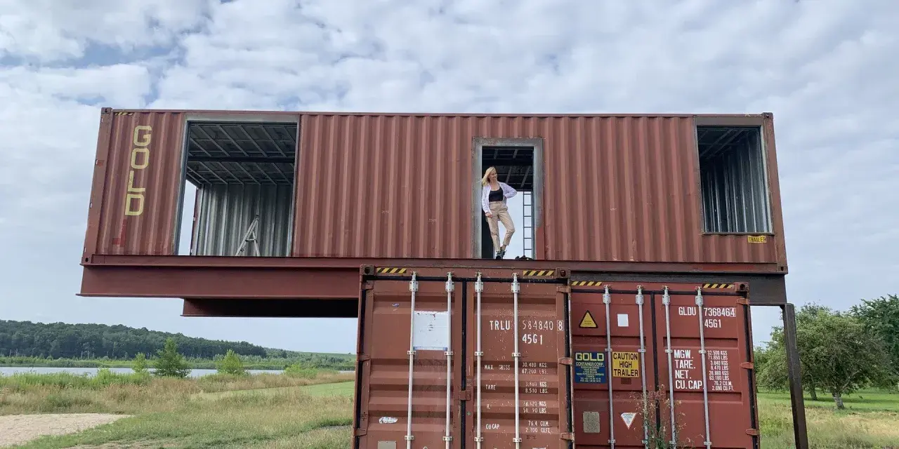 Kobieta stoi w drzwiach domu z kontenera morskiego, który jest piętrowo ułożony na innych kontenerach.