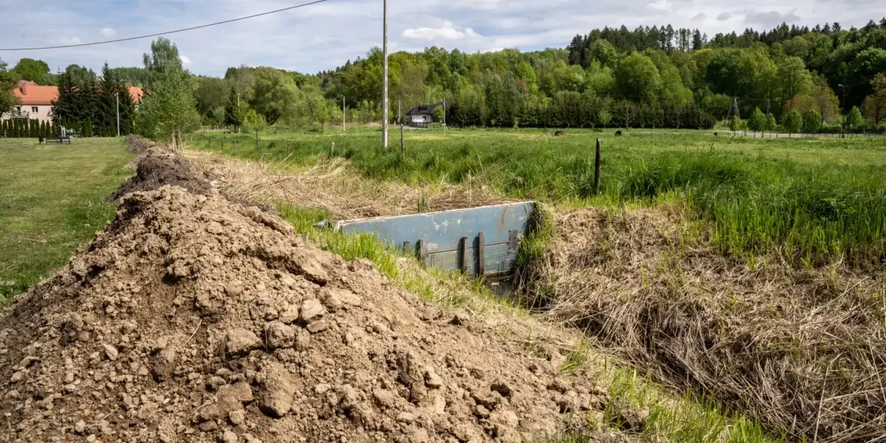 Nasyp ziemi przy rowie z niebieską przepustnicą, która może służyć do retencji wody. W tle zielone łąki i las.