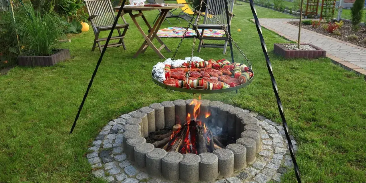 Ognisko w kamiennym kręgu, nad którym wisi okrągła taca z mięsem i warzywami na szaszłykach, gotowe do grillowania w ogrodzie.