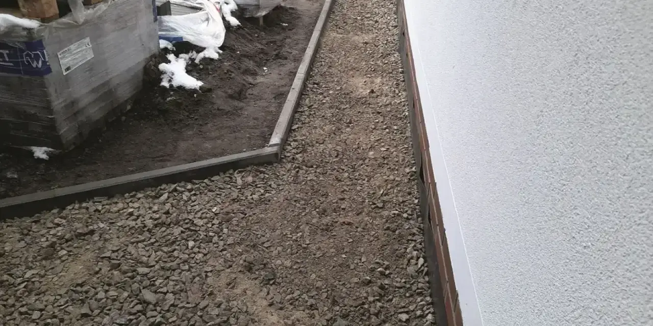 Stos **wapien** w budowie ścieżki wokół domu. Materiały budowlane leżą na ziemi, częściowo przykryte śniegiem.
