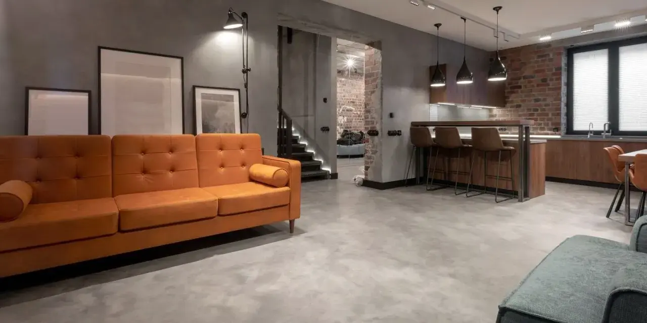 Nowoczesny salon z pomarańczową kanapą i kuchnią. Podłoga z mikrocementu na płytki nadaje industrialny charakter.
