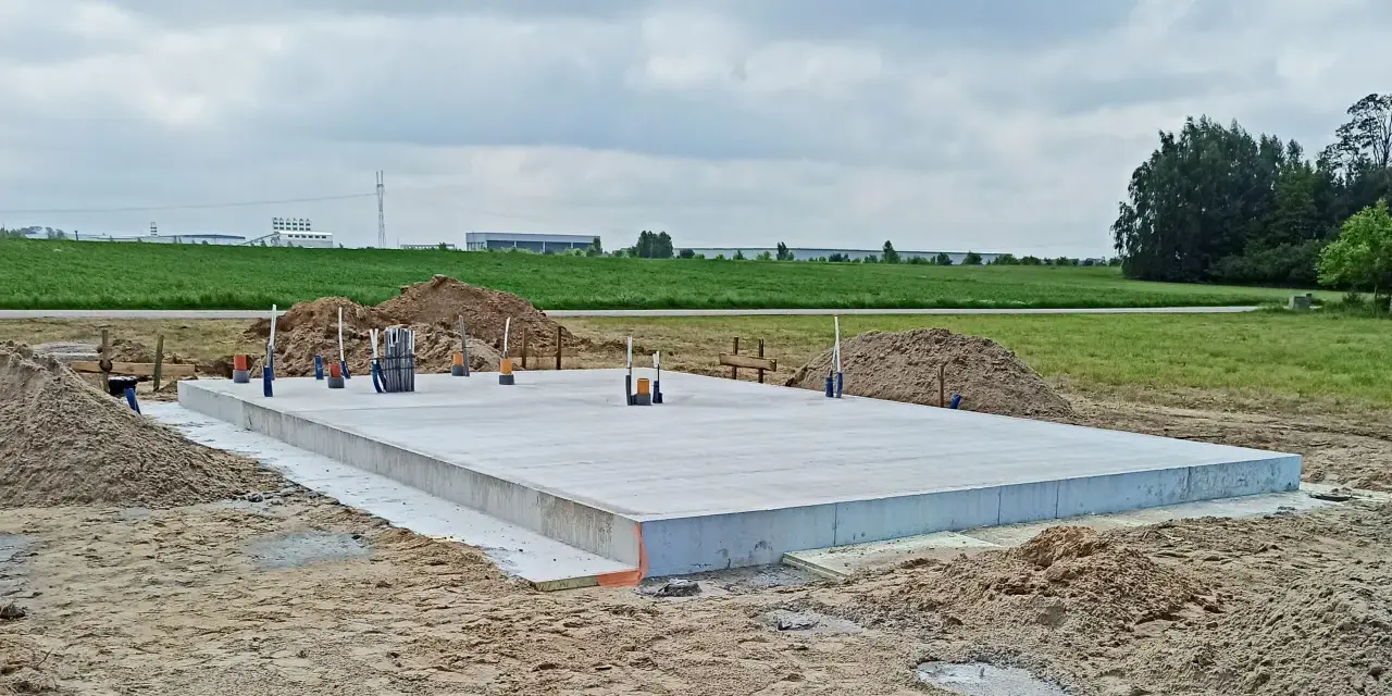 Gotowa płyta fundamentowa na budowie, otoczona piaskiem i zielenią.