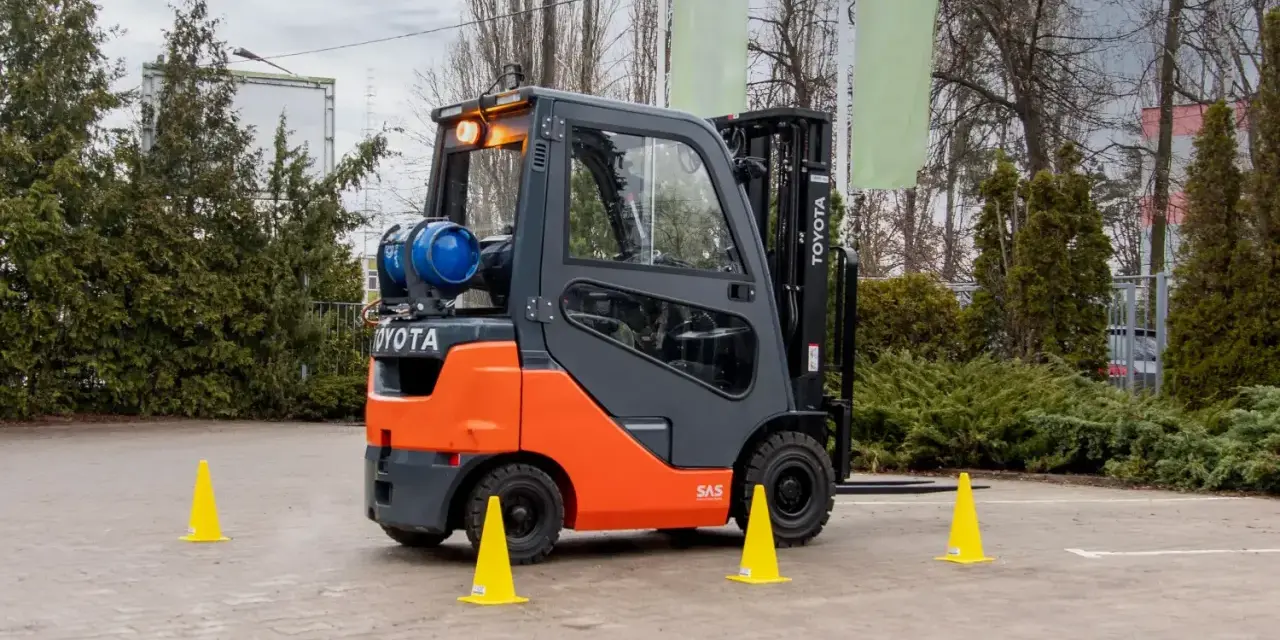 Pomarańczowo-szary wózek widłowy Toyota z butlą gazową, otoczony żółtymi pachołkami, gotowy do testów UDT.