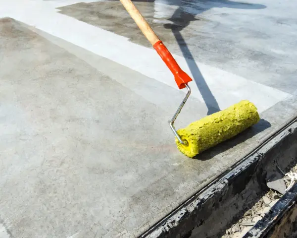 Wałek z żółtym włosiem rozprowadza środek do impregnacji betonu na szarej powierzchni.
