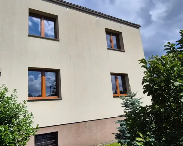 Jasny budynek z oknami o standardowych wymiarach i brązowych ramach, otoczony zielenią.