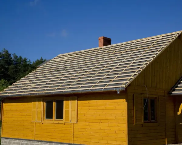 Budowa domu z żółtej deski. Na dachu układana jest papa, przygotowana pod pokrycie.