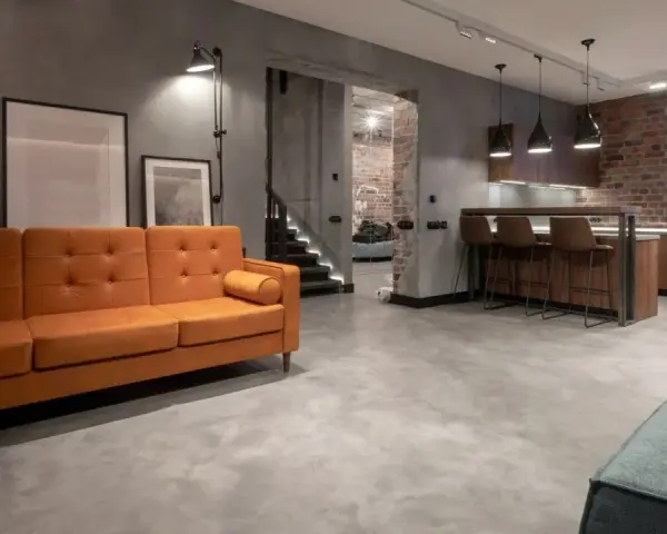 Nowoczesny salon z pomarańczową kanapą i kuchnią w stylu industrialnym. Podłoga z mikrocementu, cena za m2 tej powierzchni jest atrakcyjna.