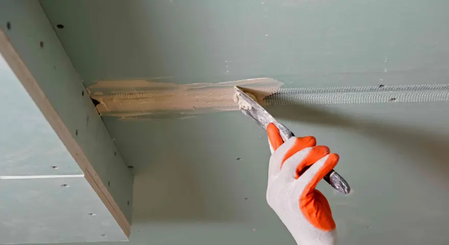 Ręka w rękawicy nakłada masę szpachlową na połączenie płyt gipsowo-kartonowych, przygotowując je do dalszych prac. Klejenie płyt GK na ściany wymaga precyzji.