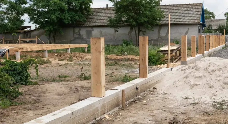 Solidny fundament pod ogrodzenie z drewnianymi słupkami, w tle budowa domu.