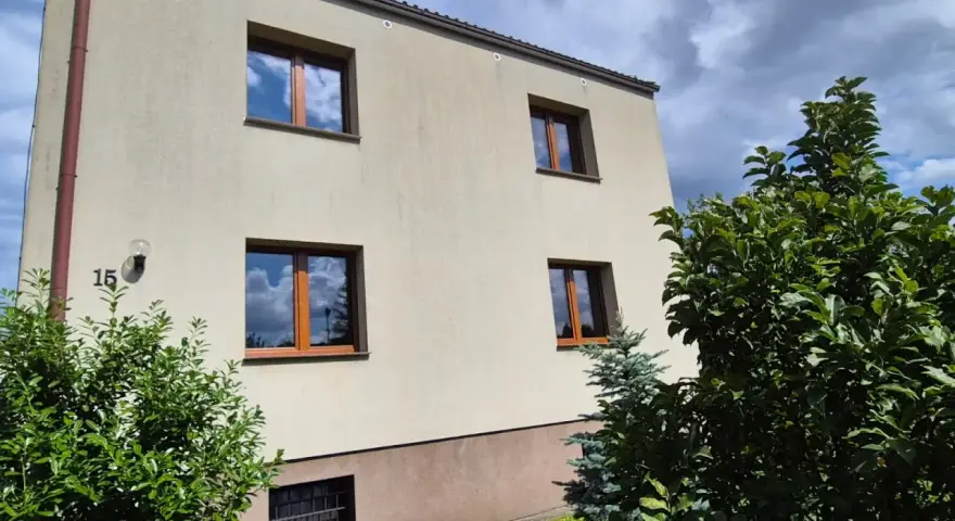 Jasny budynek z oknami o standardowych wymiarach i brązowych ramach, otoczony zielenią.