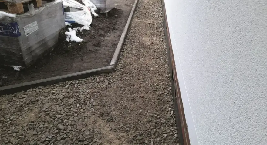 Stos **wapien** w budowie ścieżki wokół domu. Materiały budowlane leżą na ziemi, częściowo przykryte śniegiem.