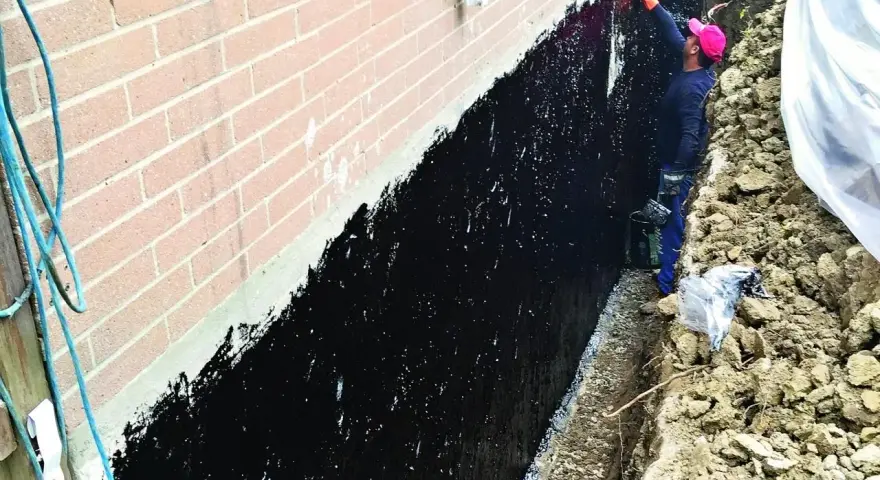 Pracownik nakłada czarną masę na ścianę fundamentową, wykonując hydroizolację.