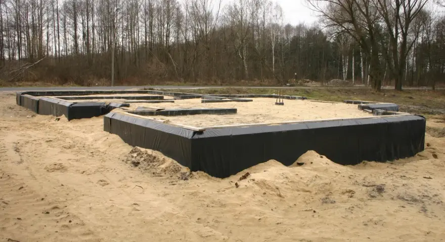 Odkopanie i izolacja fundamentów domu w piaskowym terenie. Czarna folia zabezpiecza ściany fundamentowe.