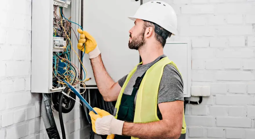 Elektryk w kasku i kamizelce odblaskowej pracuje przy skrzynce z przewodami. Dokładna wycena koszt instalacji elektrycznej.