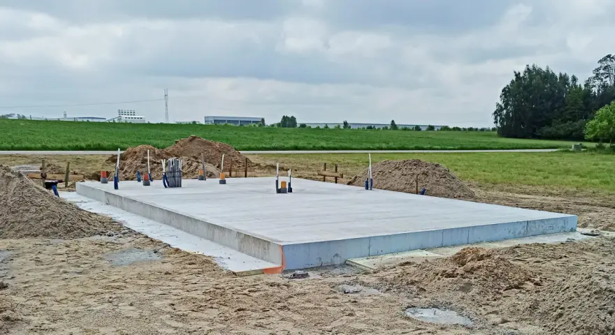 Gotowa płyta fundamentowa na budowie, otoczona piaskiem i zielenią.