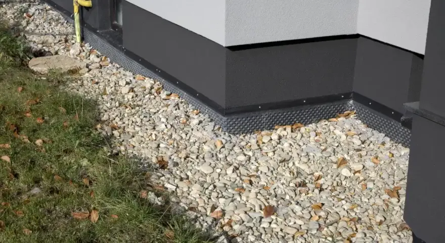 Nowy cokół na elewacji domu, otoczony kamieniami i trawą, chroni przed wilgocią.