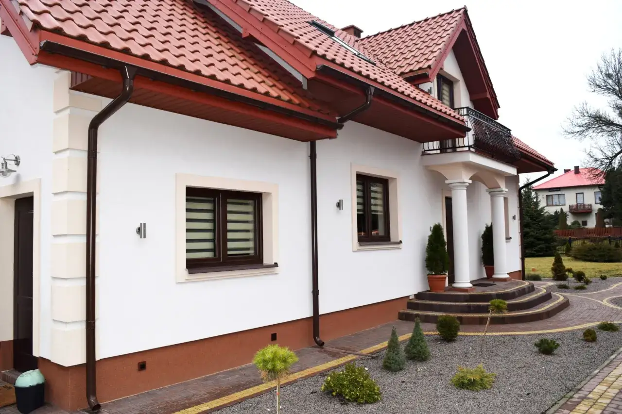 Biały dom z brązowym dachem i rynnami. Elewacja wykończona rustykalnym cokół na elewacji.