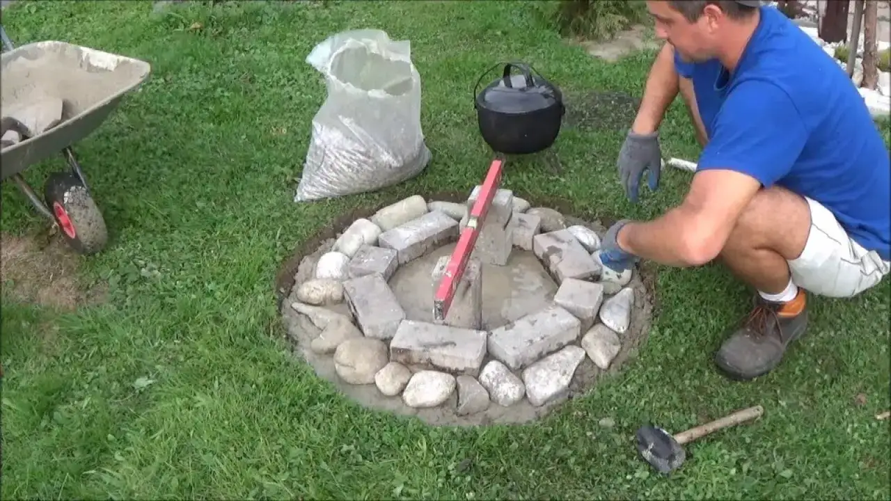 Budowa kamiennego kręgu na ognisko. Mężczyzna układa kamienie, używając poziomicy. Obok stoi wózek i worek z kruszywem.