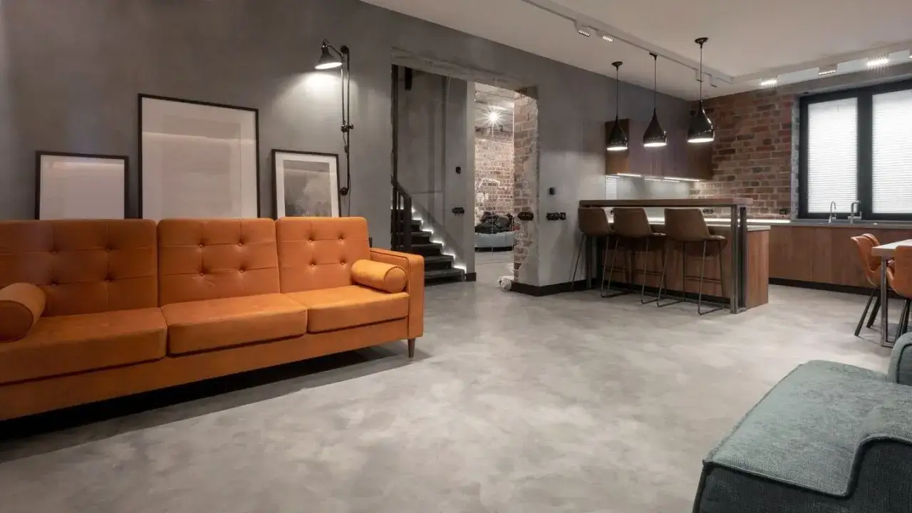 Nowoczesny salon z pomarańczową kanapą i kuchnią w stylu industrialnym. Podłoga z mikrocementu, cena za m2 tej nawierzchni jest atrakcyjna.